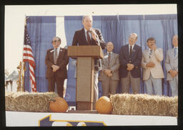 Rep. Edward Burns stands at a podium during a fall festival with pumpkins and haybale.