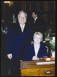 Swearing-In Day on the House Floor, undated. Rep. Thomas Tangretti stands by his desk, his wife Sandy is seated at the desk.