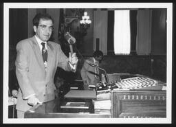 Rep. Thomas Scrimenti stands at the Speaker's rostrum holding his gavel in the air. Parliamentarian Clancy Myer stands in the background.